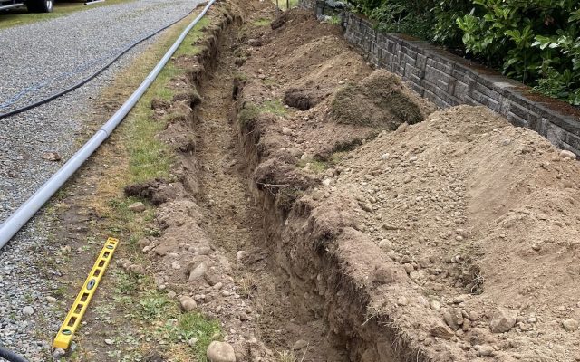 Open utility trench running alongside a gravel driveway, with grey conduit laid ready for installation and a stone block retaining wall to the right.